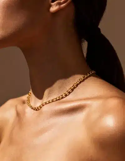 Image IA réaliste : femme portant un collier doré minimaliste sur fond neutre, lumière chaude et texture de peau naturelle.