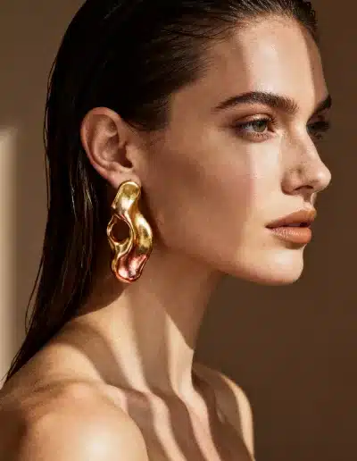 Shooting virtuel IA : femme de profil portant une boucle d’oreille dorée sculpturale, lumière dorée et esthétique éditoriale haut de gamme