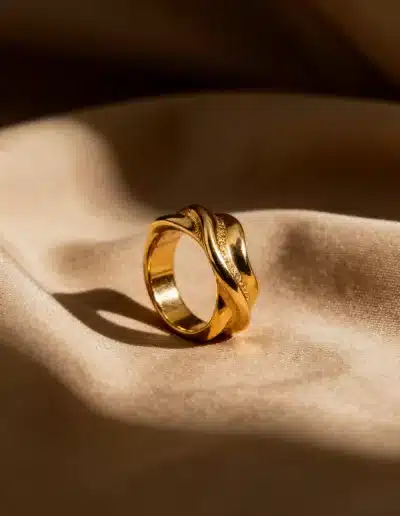 Création d’image par IA : bague dorée torsadée posée sur tissu beige satiné, lumière chaude et rendu photographique réaliste.