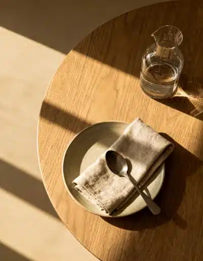 Shooting virtuel IA : table en bois clair avec assiette en grès, linge de lin et carafe d’eau, composition minimaliste et lumière matinale